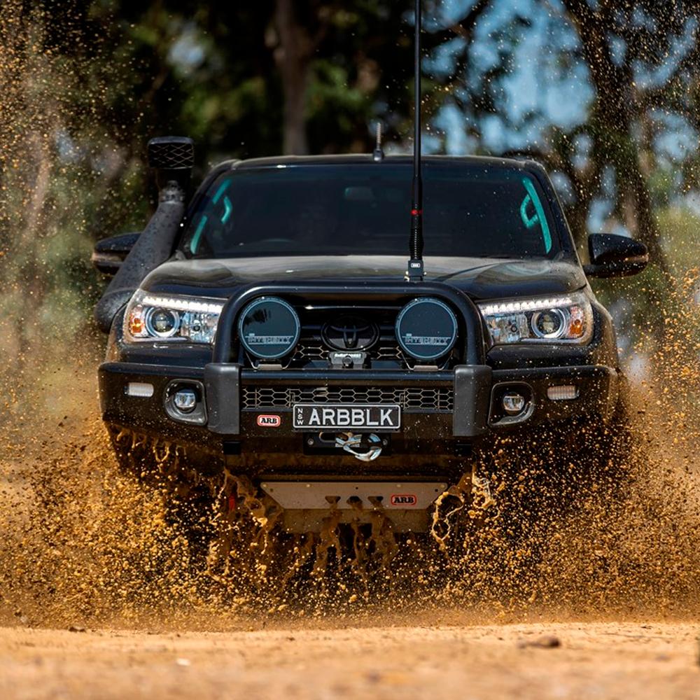 A black truck is driving through mud on a dirt road, equipped with the ARB Old Man Emu Rear Nitrocharger Sport 60080 for Toyota 4Runner/FJ Cruiser / Prado 150 Series by Old Man Emu for a smooth ride.