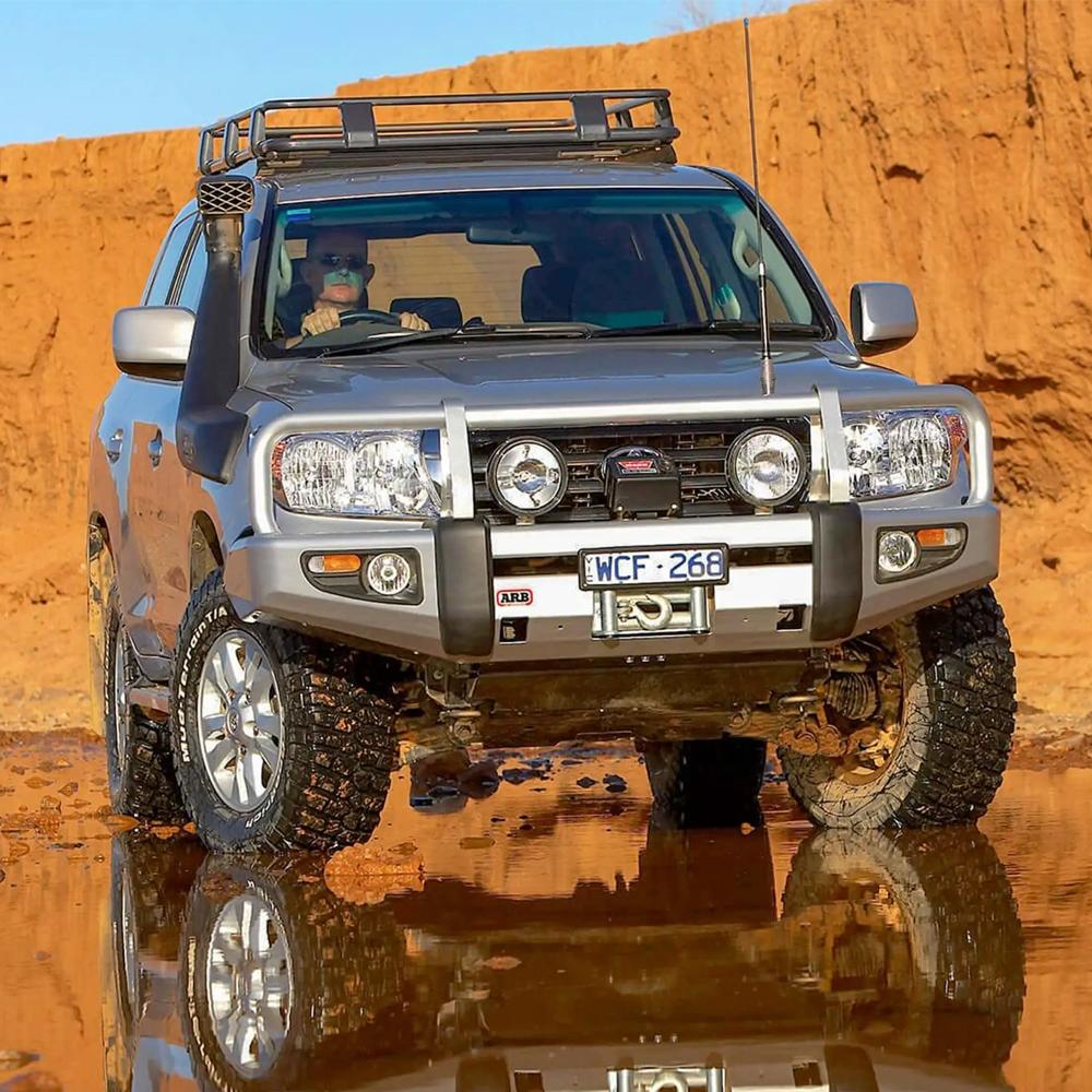 A silver Toyota Land Cruiser equipped with the ARB Roof Rack Fitting Kit 3713010 (87”x49”) for Toyota Landcruiser 100/105 Series and Lexus Lx470 navigating through a high capacity muddy terrain, ensuring secure storage for all belongings.