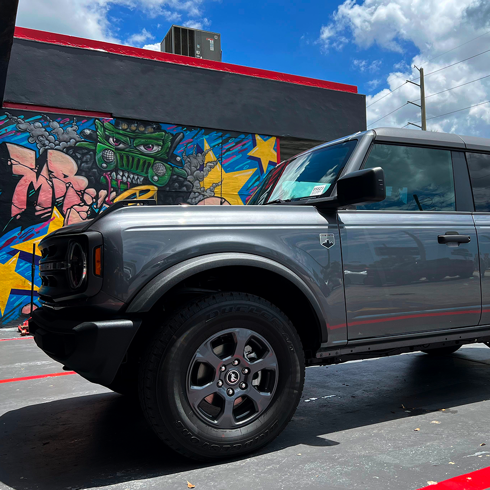 A gray Ford Bronco parked in front of a graffiti wall with an installation vibe, equipped with Old Man Emu ARB Front Coil Springs 3198 for Bronco Base, Big Bend, Outer Banks, Wildtrack (2021-2022) - Light Loads Estimated Lift.