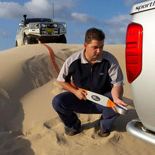Load image into Gallery viewer, A man performing vehicle recovery in front of a truck stuck on a sand dune, using an ARB715LB Snatch Block Strap from ARB.