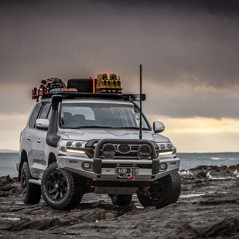 An ARB Base Rack Deflector For Toyota Land Cruiser 200 Series (2008-2022) is parked on a rocky beach.