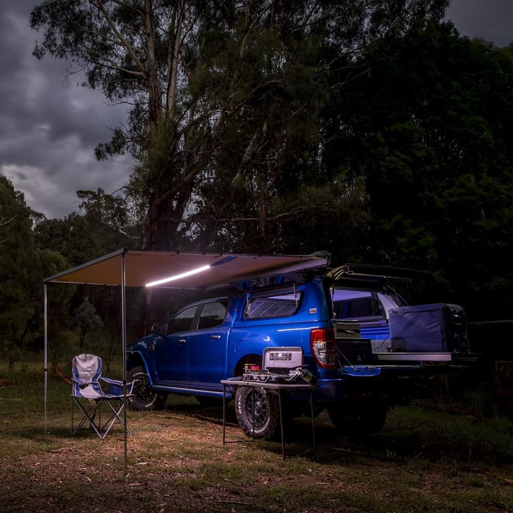 A waterproof Toyota pickup truck with roof racks and ARB retractable awnings, equipped with a camping table and chairs.