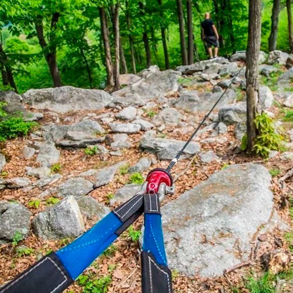 A person is using a Factor 55 Shackle Mount in Red 00015-01 to hold a rope on a rock in the woods.