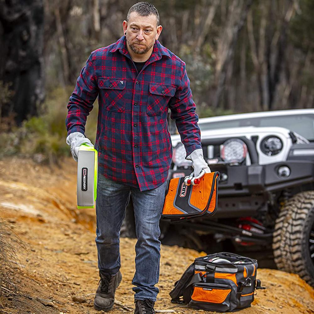 A man wearing a plaid shirt carefully navigates down a dirt road with a jeep, equipped with the ARB Orange Large Recovery Bag ARB501A.