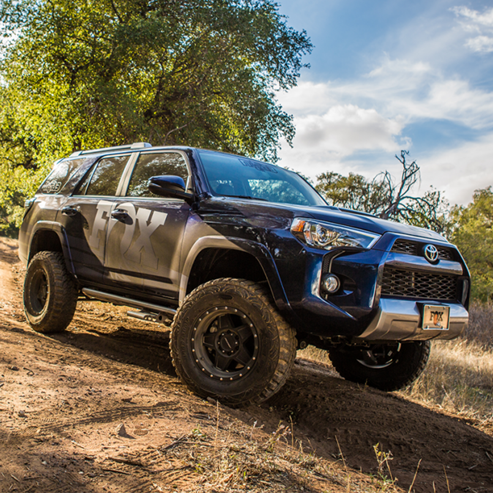 A blue truck or SUV is driving down a dirt road equipped with Fox Racing Performance Series 2.0 IFP smooth body shocks featuring advanced shock technology, specifically the FOX 2.0 Performance Series Smooth Body IFP - Rear Shock 980-24-679 for Toyota 4Runner / FJ Cruiser / Land Cruiser Prado120 & 150.
