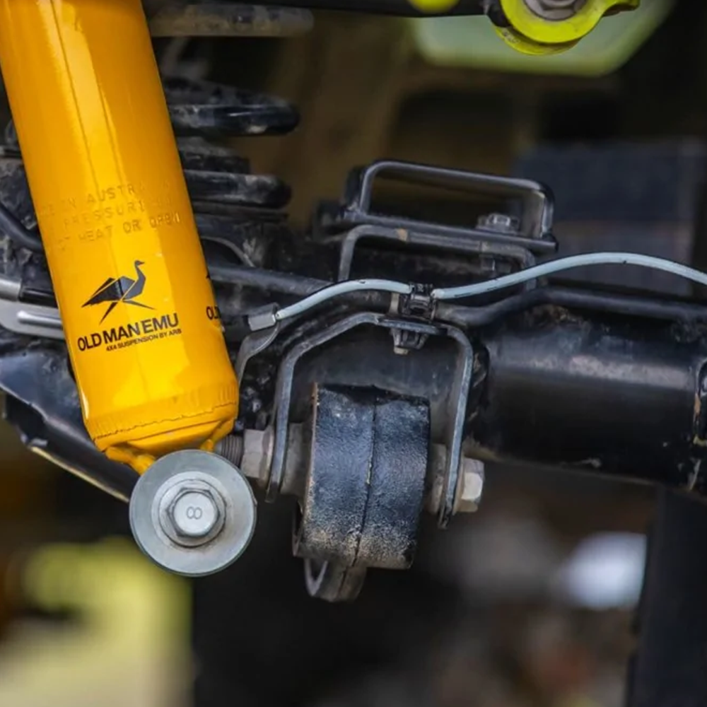 A close up of a vehicle's suspension featuring a yellow Old Man Emu Caster Correction OMECA001 for Suzuki Jimny (2018-2021) 4th GEN - CYL. 1.5L PETROL ENGINE caster correction kit for improved wheel alignment and responsive steering.