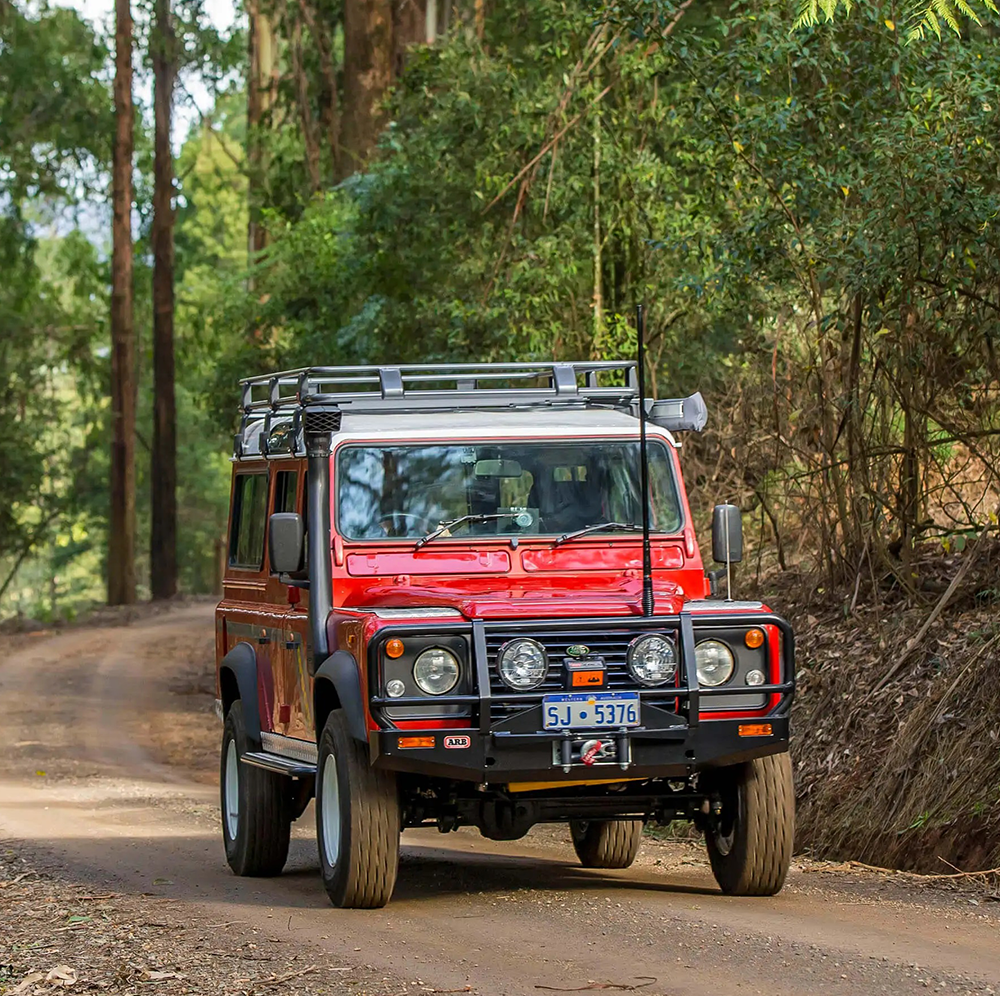 A red Old Man Emu Land Rover with increased ride height is driving down a dirt road.