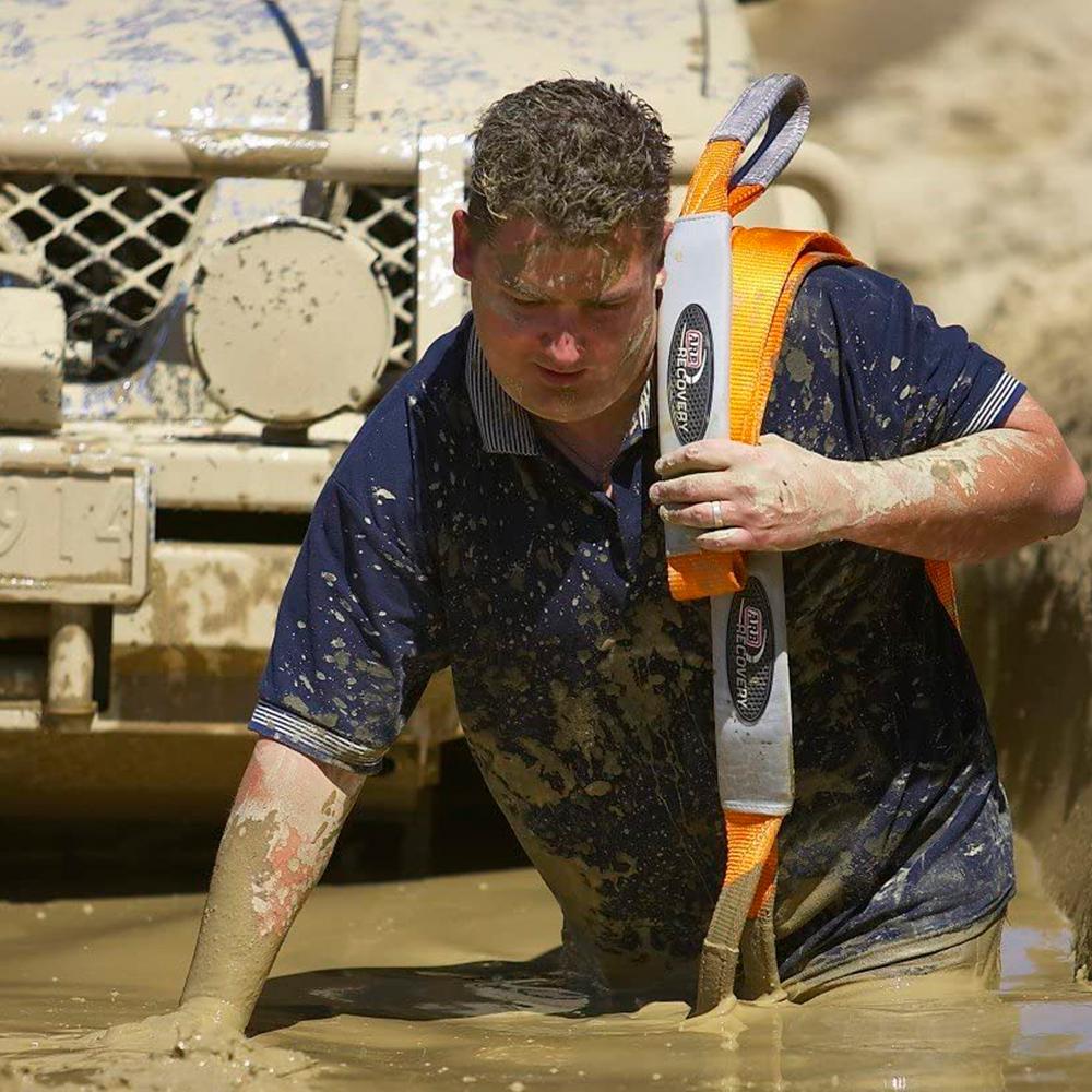 The man, wearing a blue shirt, is demonstrating vehicle recovery using the Snatch Block Strap ARB715LB from the brand ARB.