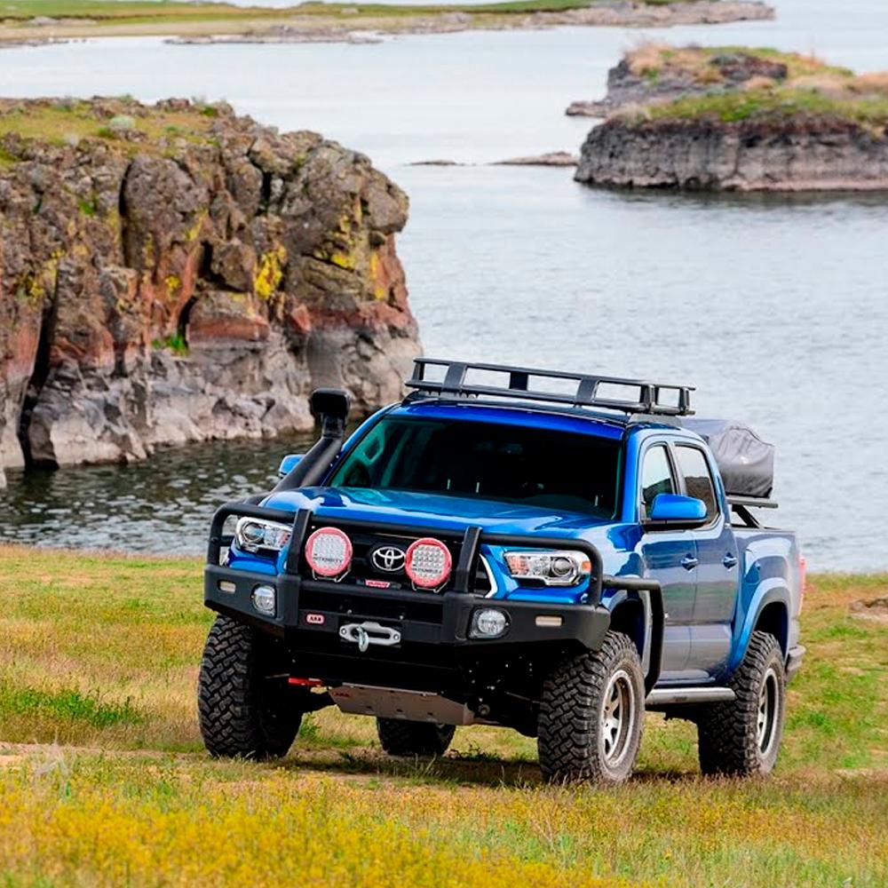 A blue Old Man Emu Toyota Tacoma with a suspension lift is parked in front of a lake.