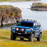 A blue Old Man Emu Toyota Tacoma with a suspension lift is parked in front of a lake.