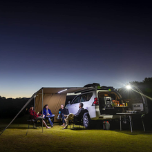 Load image into Gallery viewer, A group of people sitting around a camper van with ARB Touring Awning with Light 814409 at night.